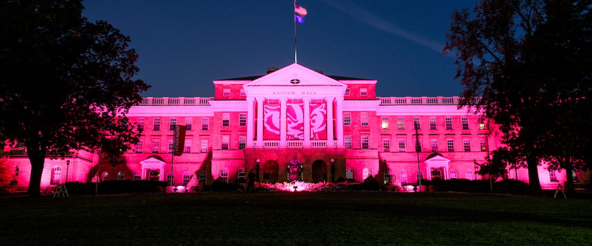 UW–Madison’s Bascom Hall lit in pink lights at night with a large Bucky Badger banner