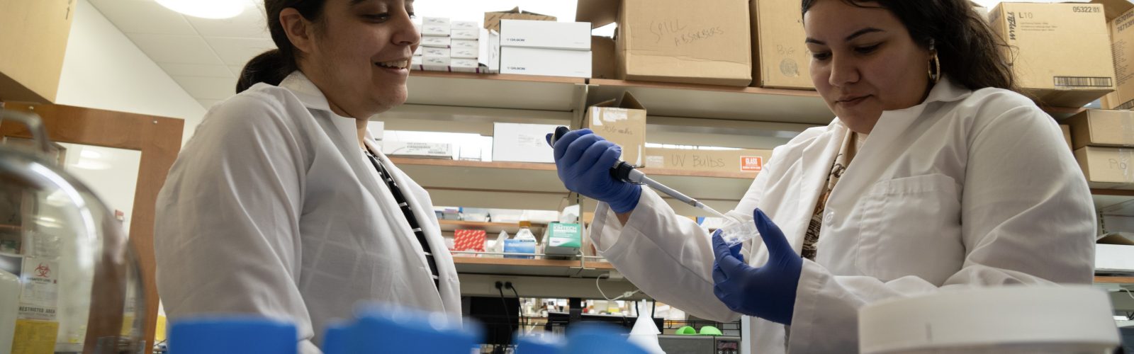 Technician demonstrates pipette use to colleague at lab bench with containers and supplies